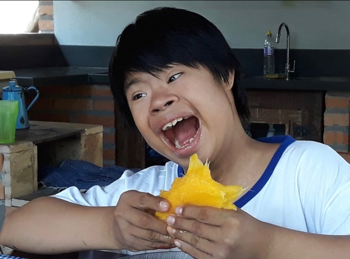 Menino indígena com síndrome de Down e surdez, de cabelos pretos, blusa branca comendo manga