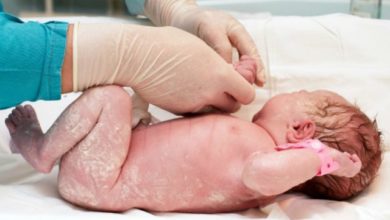 Bebê branco recém nascido, deitado em pano branco sendo examinado por médico com luvas cirúrgicas e jaleco verde agua