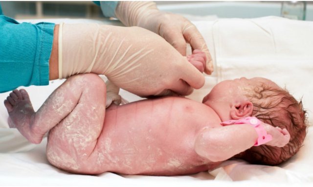 Bebê branco recém nascido, deitado em pano branco sendo examinado por médico com luvas cirúrgicas e jaleco verde agua