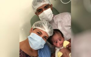 Homem e mulher branco de mascara e touca cirúrgica, eles estão com a filha bebê menina que nasceu com síndrome rara 