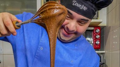 Jovem com síndrome de down branco na cozinha, ele veste um avanetal azul, e um chapeu de cozinheiro preto e esta com um fuet com massa para bolo de chocolate