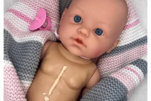 Boneca bebê, branca de olhos azul enrolada em uma manta de lã listada, rosa, cinza e pink, a boneca tem uma chupeta e tem uma cicatriz no peito