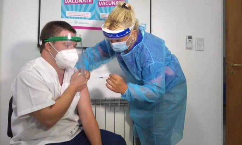 Imagem de funco branco com um quadro de funddo azl, uma enfermeira loira com avental azul e mascara vacinando um homem com síndrome de down branco que veste uma camiseta branca e calça preta
