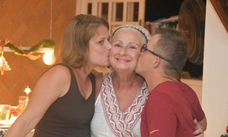 Uma mae com seus dois filhos, a jovem está a sua direita e o jovem a sua esquerda eles estão de perfil beijando a face da mãe