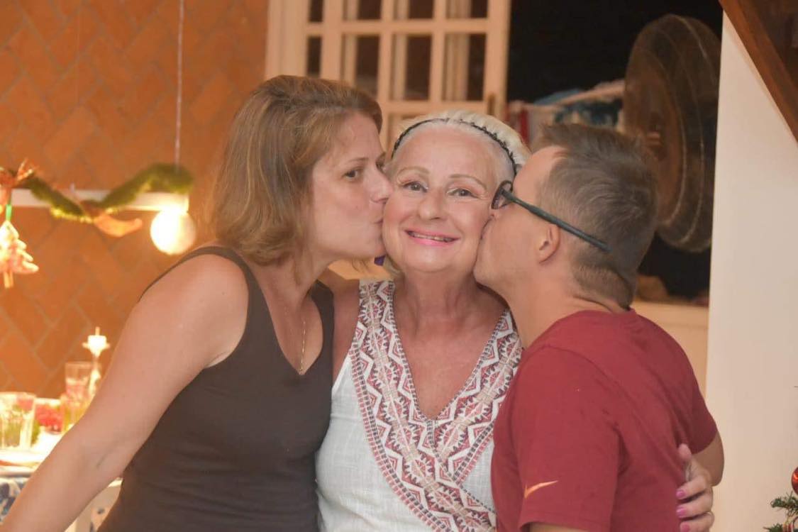 Uma mae com seus dois filhos, a jovem está a sua direita e o jovem a sua esquerda eles estão de perfil beijando a face da mãe