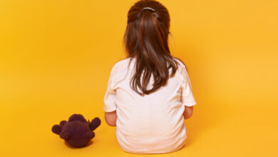 Imagem de fundo amarelo com uma menina pequena sentada de costas com um urso marrom ao seu lado . ela veste uma camiseta branca e tem cabelos castanhos presos em meio rabo