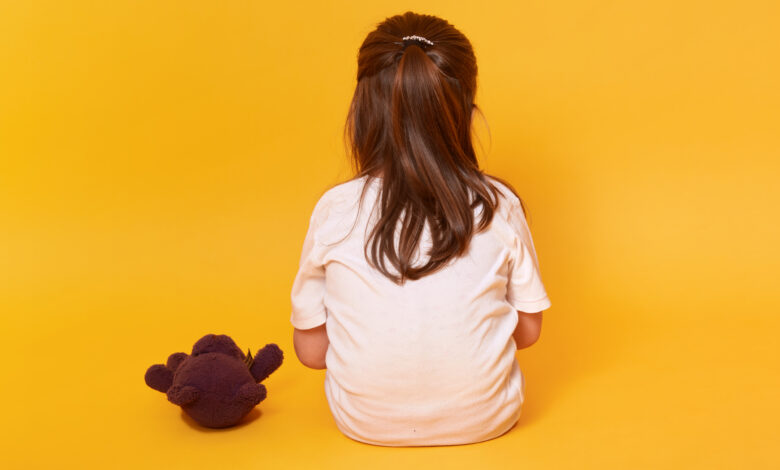 Imagem de fundo amarelo com uma menina pequena sentada de costas com um urso marrom ao seu lado . ela veste uma camiseta branca e tem cabelos castanhos presos em meio rabo