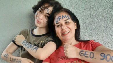 Duas mulheres brancas de cabelos castranho, a da direita é a filha de cabelos curtos, camiseta marrom e com escritos em caneta azul nos braços e no rosto UFMS 2º lugar farmácia, e do lado direito a mãe de cabelos longos, blusa vermelha e escritos em caneta azul no rosto e nos braços UPMS 4º lugar geografia