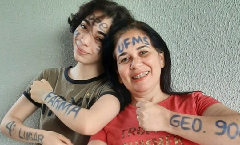 Duas mulheres brancas de cabelos castranho, a da direita é a filha de cabelos curtos, camiseta marrom e com escritos em caneta azul nos braços e no rosto UFMS 2º lugar farmácia, e do lado direito a mãe de cabelos longos, blusa vermelha e escritos em caneta azul no rosto e nos braços UPMS 4º lugar geografia