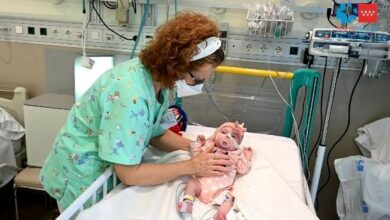 Bebe menina deitada em uma maca de hospital vestindo uma roupa rosa e um laço, conectada a dispositivos que a ajudam a viver, com uma enfermeira ao seu lado com avental verde colocando a mão sobre o bebê como sinal de cuidado