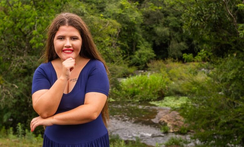 imagem com fundo de plantas há uma jovem branca de cabelos e olhos castanhos, com vestido azul, sua mão esquerda apoia seu queixo e a direita sua cintura