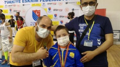 imagem com fundo branco com escritos sobre o campeonato de esgrima, há 3 pessoas na foto, um homem branco calvo de blusa amarela, uma criança com uniforme azul e uma medalha e ouro e um homem com a camiseta do evento e um cracha