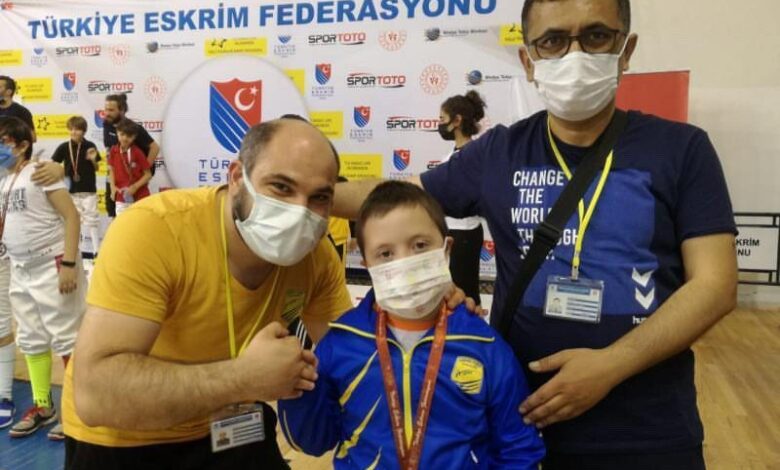 imagem com fundo branco com escritos sobre o campeonato de esgrima, há 3 pessoas na foto, um homem branco calvo de blusa amarela, uma criança com uniforme azul e uma medalha e ouro e um homem com a camiseta do evento e um cracha