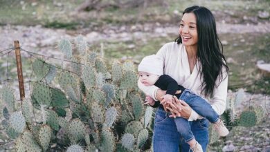 Foto: imagem ao fundo deserto com cactos, ha uma mãe de origem asiática, ela tem cabelos longos negros e segura seu bebê de gorro branco, blusa azul e calca jeans