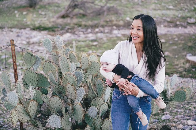 Foto: imagem ao fundo deserto com cactos, ha uma mãe de origem asiática, ela tem cabelos longos negros e segura seu bebê de gorro branco, blusa azul e calca jeans
