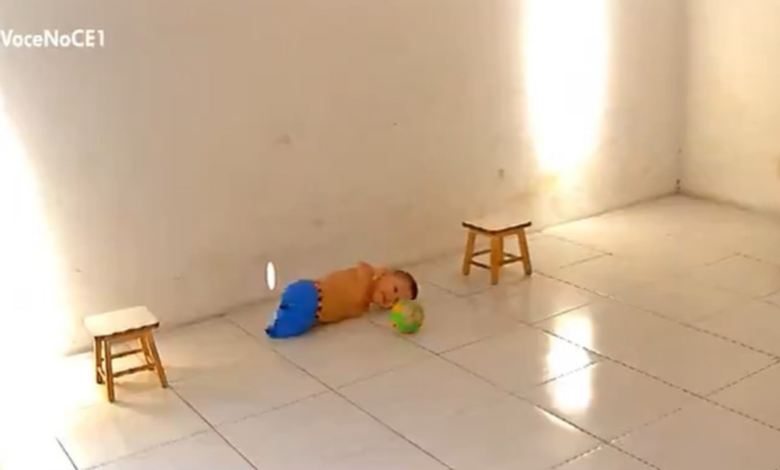 Miguel usa short azul, está deitado no chão brincando com uma bola de futebol.