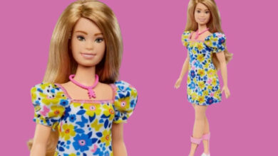 Imagem de fundo rosa com uma boneca Barbie com síndrome de Down em pé, vestida com um vestido florido azul e amarelo.