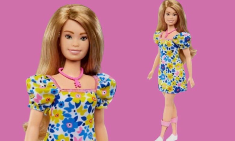 Imagem de fundo rosa com uma boneca Barbie com síndrome de Down em pé, vestida com um vestido florido azul e amarelo.