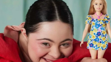 Imagem de fundo verde com uma jovem com síndrome de down com os cabelos castanhos presos, com uma blusa vermelha sorrindo, segurando uma barbie com síndrome de down com vestido florido