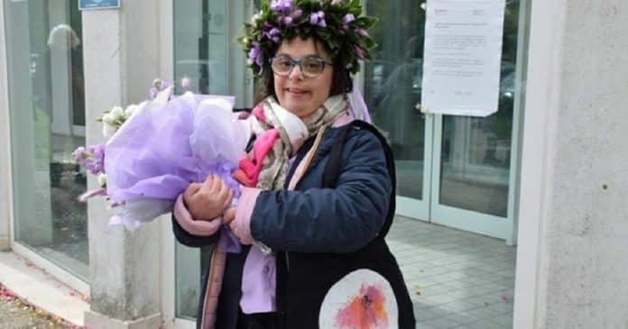 Imagem de uma jovem branca com síndrome de de down, de óculos, com casaco azul, carregando um ramalhete de flores diante de uma porta de vidro