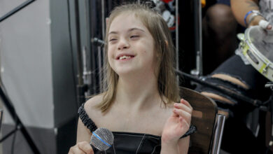 Menina branca loira com síndrome de Down com uma blusa preta segurando um microfone sorrindo