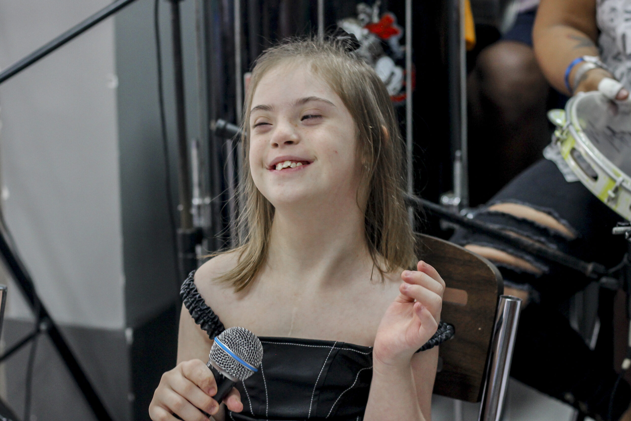 Menina branca loira com síndrome de Down com uma blusa preta segurando um microfone sorrindo