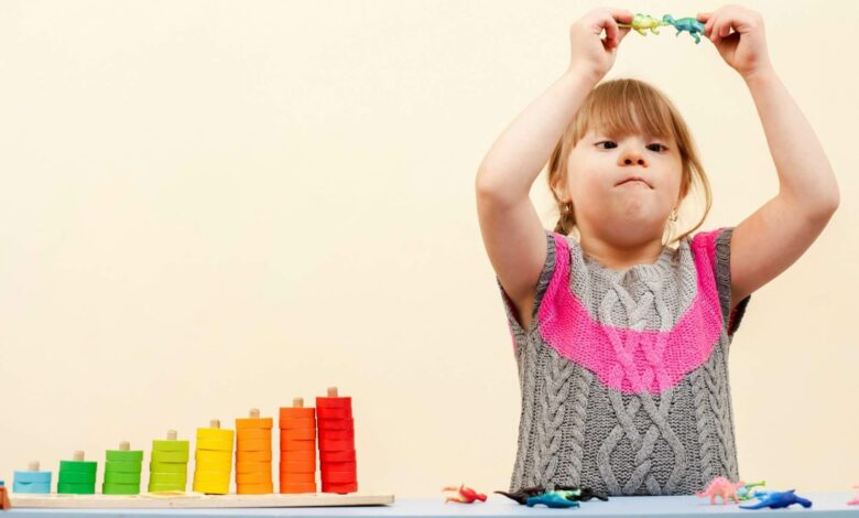 Imagem de fundo creme, no canto direito uma menina branca com síndrome de down, cabelos loiros com franja segurando um brinquedo no alto e ao lado dela um brinquedo colorido de encaixar